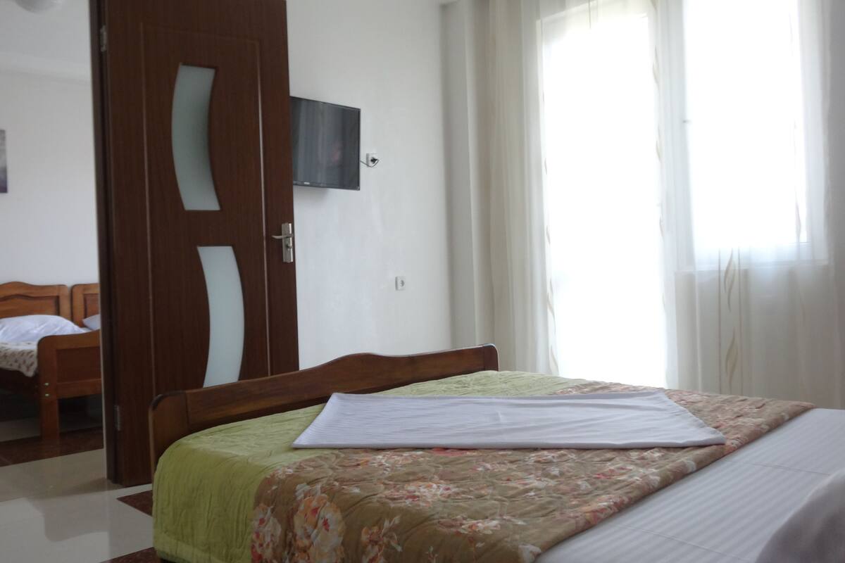 A hotel room with a bed, a wooden wardrobe, a television on the wall, and a door leading to another room.