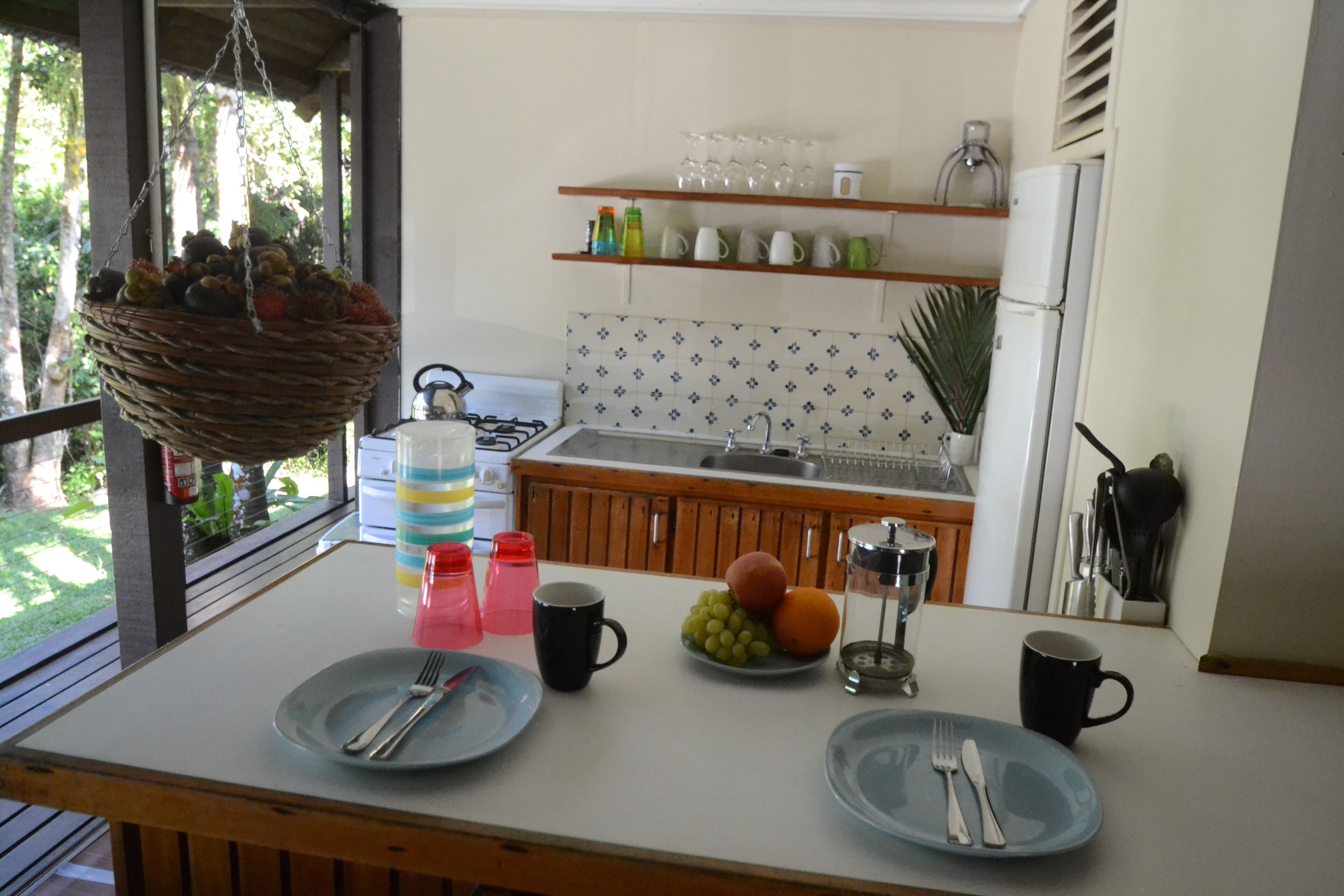 A kitchen with a dining table set for two, a hanging fruit basket, and a variety of kitchen utensils and appliances.
