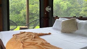 Una cama con sábana blanca y manta color beige, cabecera de madera y una mesita de noche con almohada blanca.