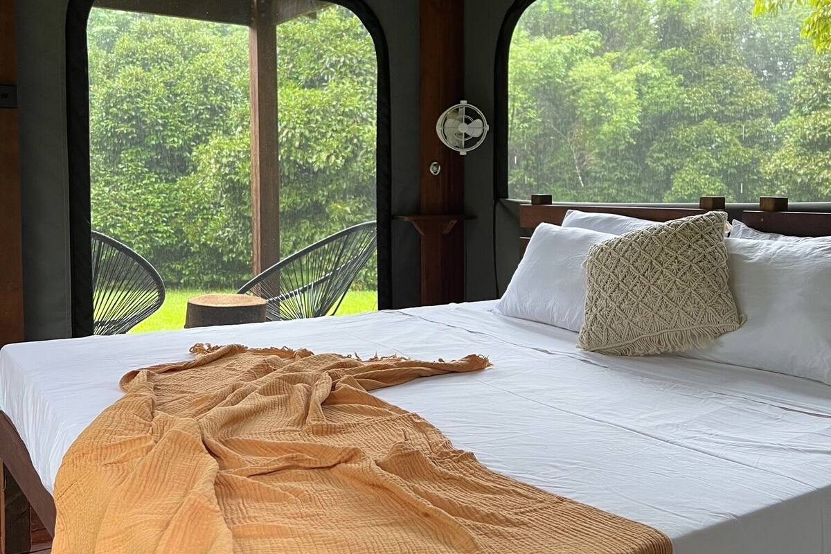 Un lit avec un drap blanc et une couverture beige, une tête de lit en bois et une petite table de chevet sur laquelle repose un oreiller blanc.