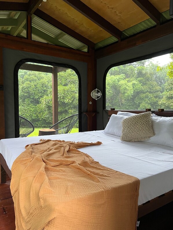 Un lit avec un drap blanc et une couverture beige, une tête de lit en bois et une petite table de chevet sur laquelle repose un oreiller blanc.