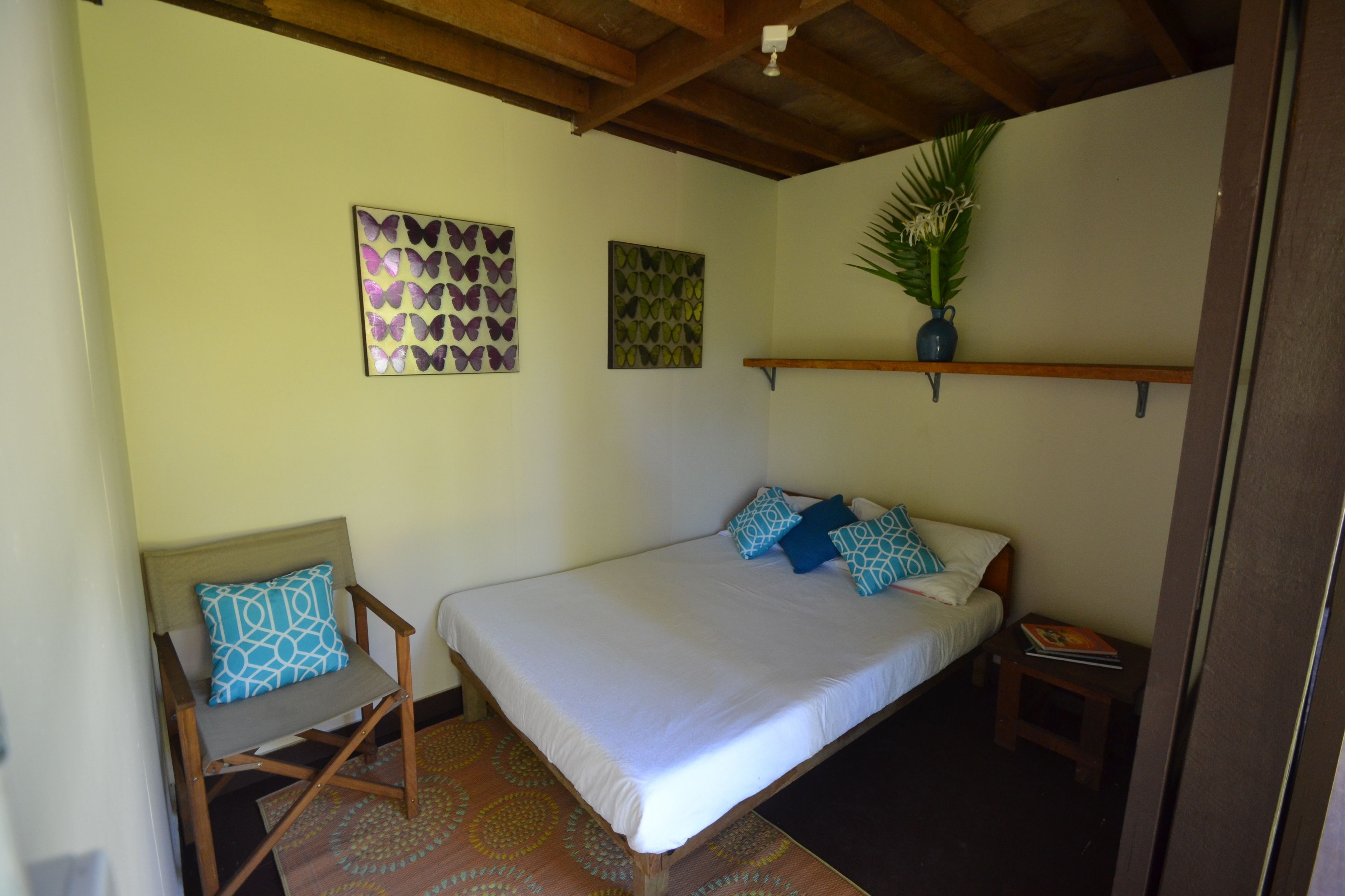 Quarto com uma cama, uma cadeira, uma estante com uma planta e duas obras de arte nas paredes com padrão de borboletas.