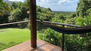 Una hamaca en una terraza de madera con vistas a un jardín exuberante y montañas lejanas.