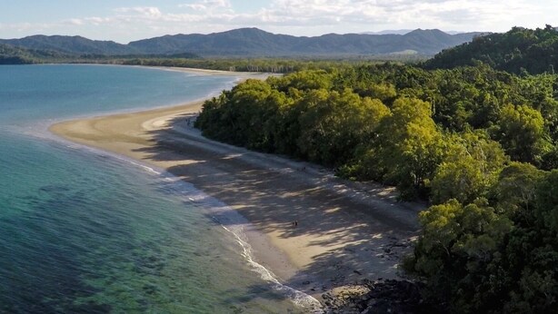 Un paisaje costero con playa, aguas cristalinas y colinas boscosas circundantes.