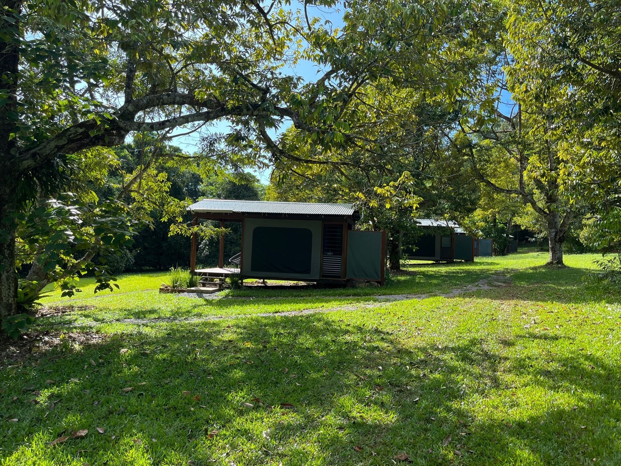 A campsite with two small, open-sided shelters with metal roofs, surrounded by trees and grass.