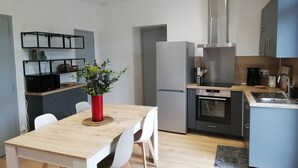 A modern kitchen with a dining area, featuring a wooden table, white chairs, a red vase with flowers, and stainless steel appliances.