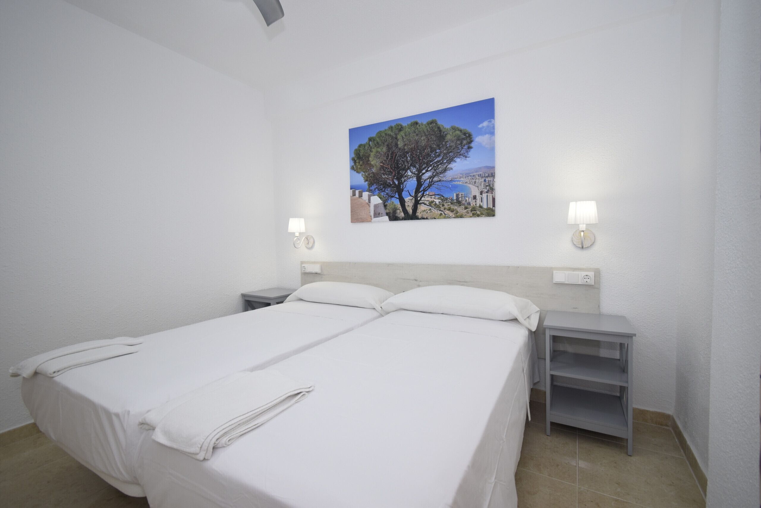 A double bed with white linens, two bedside tables, a wall-mounted lamp, and a painting of a tree and landscape.