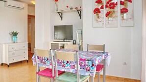 Ein Essbereich mit Tisch, bedeckt mit einer bunten Tischdecke, Stühle mit rosa Kissen, einem Fernseher auf einem Schrank und einem Regal mit Dekorationen.