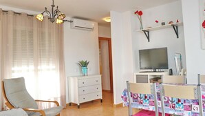 Ein Wohnzimmer mit Fernseher, einem Esstisch mit bunten Tischdecken, einem Sessel, einer Couch, einer Topfpflanze und einer Deckenleuchte.