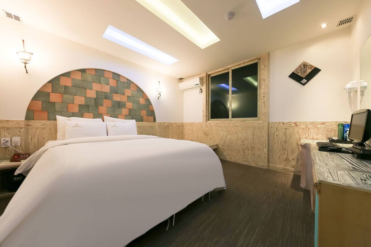 A hotel room with a bed, a desk, a chair, a window, and a wall-mounted air conditioning unit.
