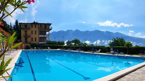 A swimming pool with clear blue water, surrounded by lounge chairs and a building with a mountain view in the background. - Appartamento 105 Benaco (Tignale)