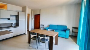 A modern kitchen with white cabinets and a coffee maker, a wooden dining table with chairs, and a bright blue sofa in a living area. - Appartamento 105 Benaco (Tignale)