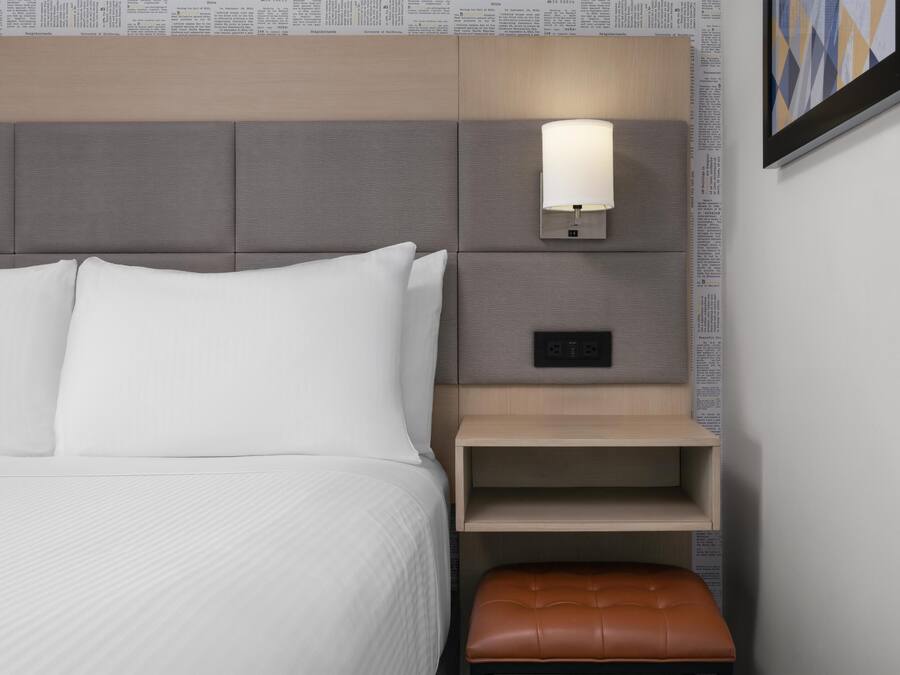 A neatly made bed with white linens, a bedside table with a lamp, and a brown leather bench.