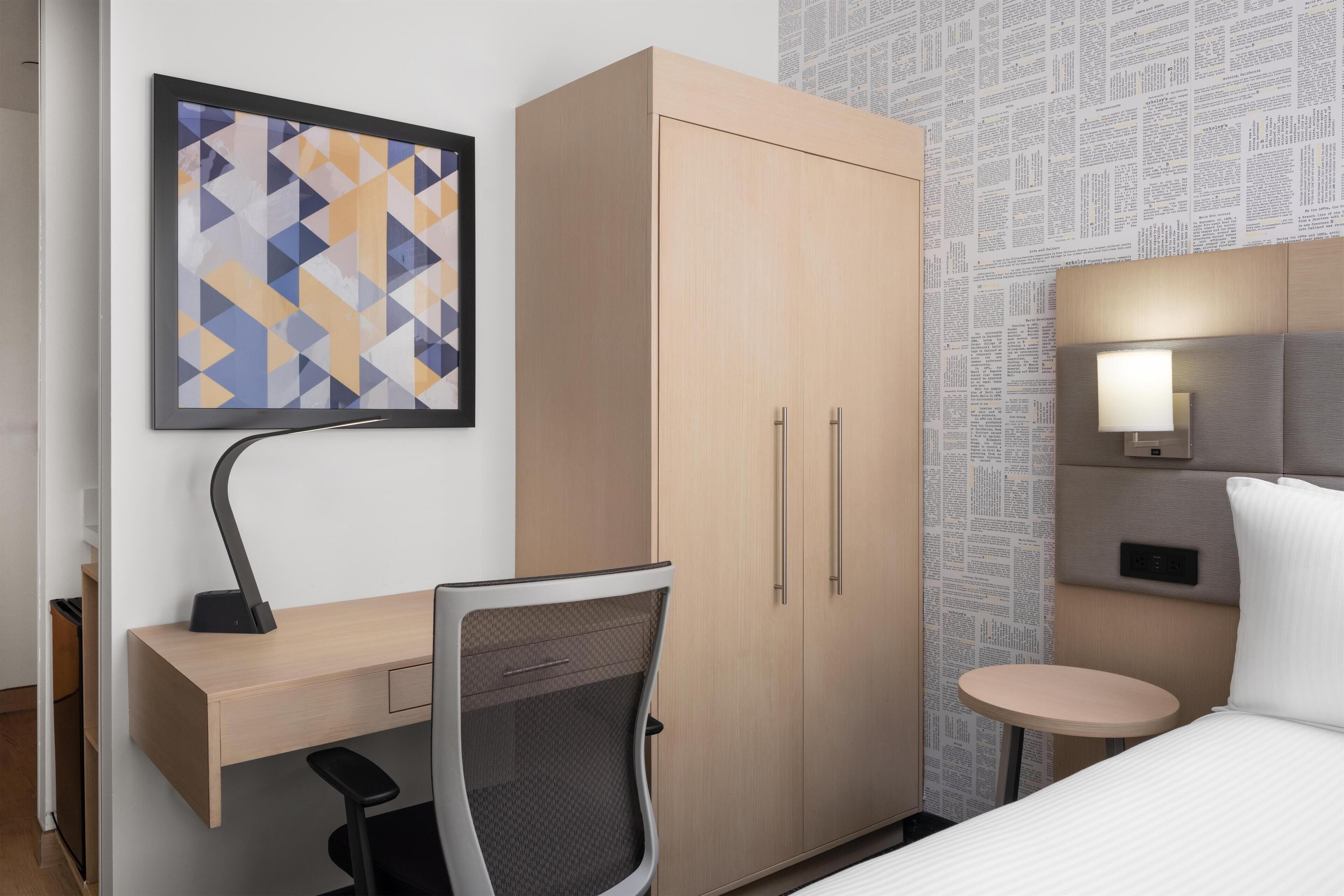 Une chambre d’hôtel avec un bureau en bois, une chaise, un lit, une petite table ronde, une lampe fixée au mur et une œuvre d’art abstraite encadrée.