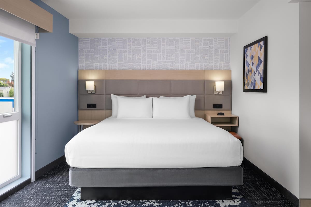 A hotel room with a large bed, two bedside tables, a headboard with integrated lighting, a window with a view, and a patterned wall.