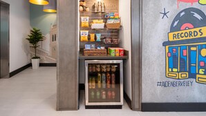Un mini refrigerador con bebidas, un estante con botanas como papas fritas y otros aperitivos, y un mural con un tocadiscos y el texto '#AIDENBERKELEY'.
