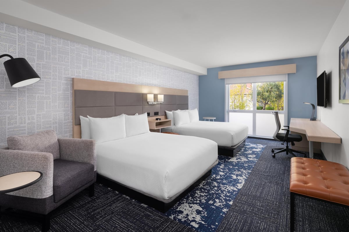 A hotel room with two beds, a desk, a chair, a sofa, and a lamp.