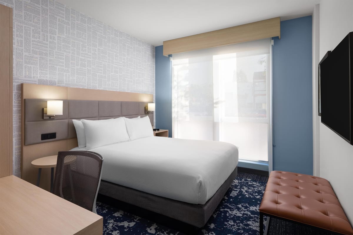 A hotel room with a large bed, a desk, a chair, a TV, and a window with a view.