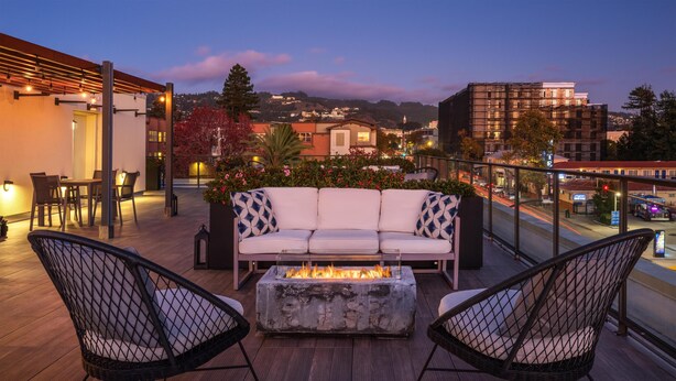 Una terraza en la azotea con un sofá blanco, dos sillas de mimbre, un hogar para fuego y vistas de la ciudad al atardecer.