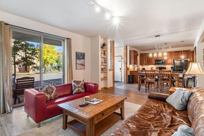 Un salón amplio con un sofá de cuero rojo, una mesa de centro de madera y un sofá de cuero marrón. Se ve una cocina con armarios de madera y un comedor con una mesa y sillas al fondo.