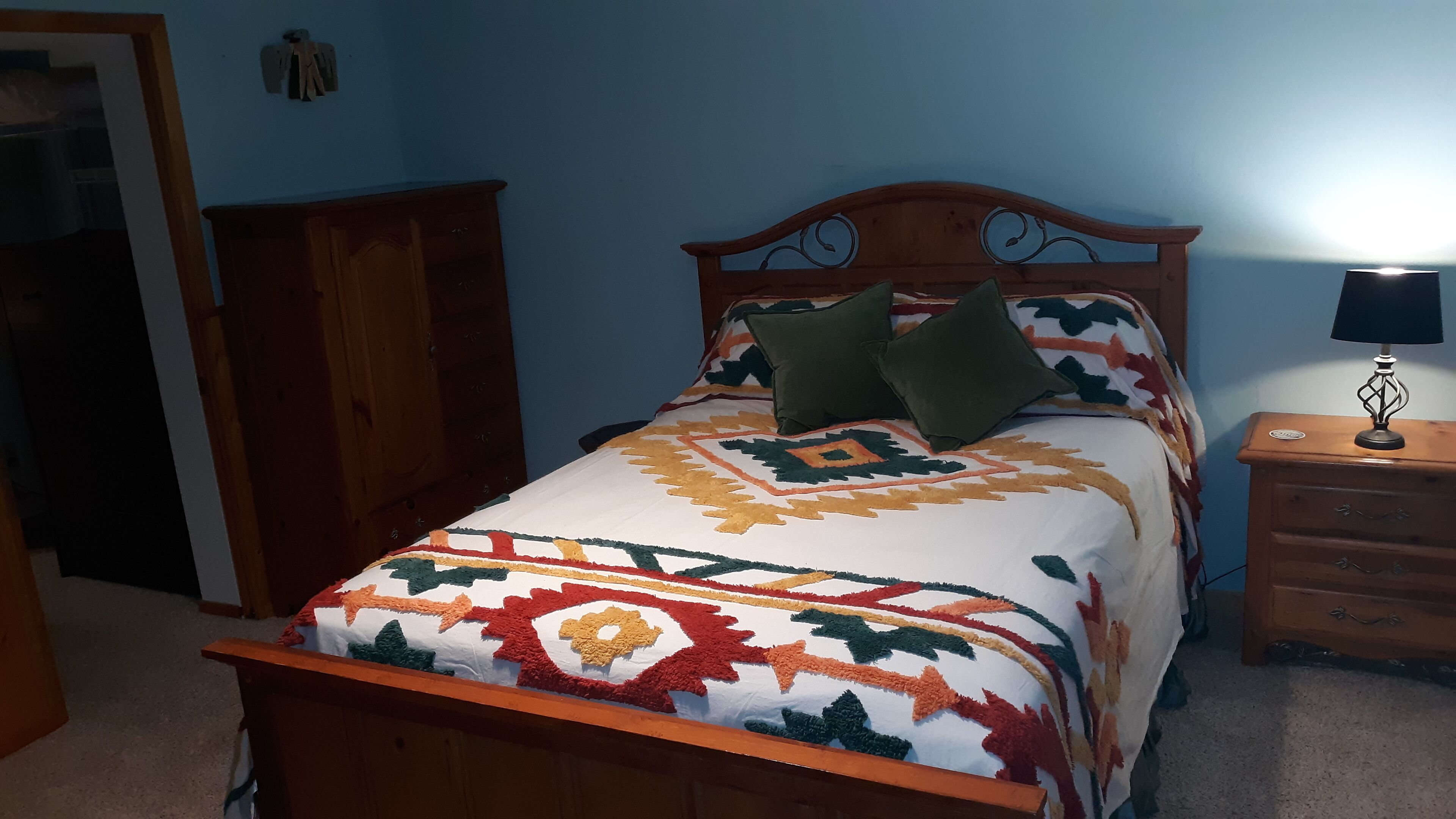 A bed with a patterned bedspread, a wooden headboard, two pillows, a nightstand with a lamp, and a wooden wardrobe.