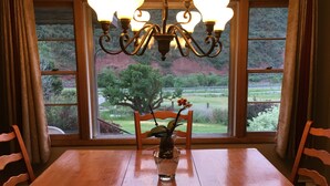 Une table à manger avec des chaises, un lustre et une vue sur un jardin, visible à travers de grandes fenêtres.