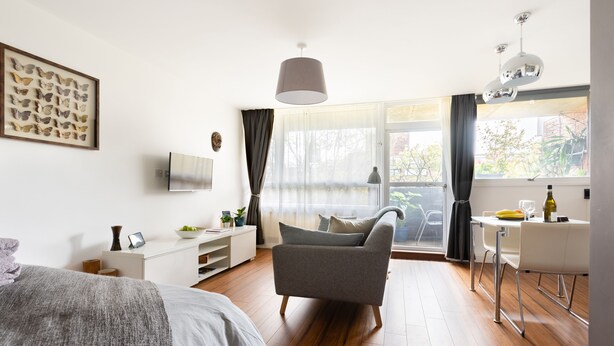 A modern bedroom with a bed, a grey armchair, a desk with a chair, a TV, and a large window with curtains. - Smart Modern Studio in Pimlico with Garden Views (London)