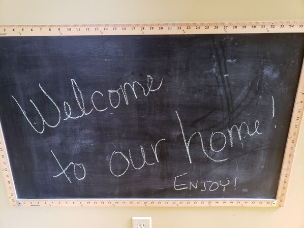 A blackboard with "Welcome to our home! ENJOY!" written on it, mounted on a wall with a measuring tape beside it.