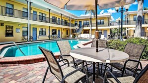 Uma área de piscina de hotel com mesa e cadeiras sob um guarda-sol, rodeada por varandas e um céu azul límpido.