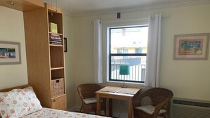 Um quarto com cama, uma estante de madeira, duas cadeiras de vime, uma mesinha, uma janela com cortinas e um ar condicionado fixado na parede.