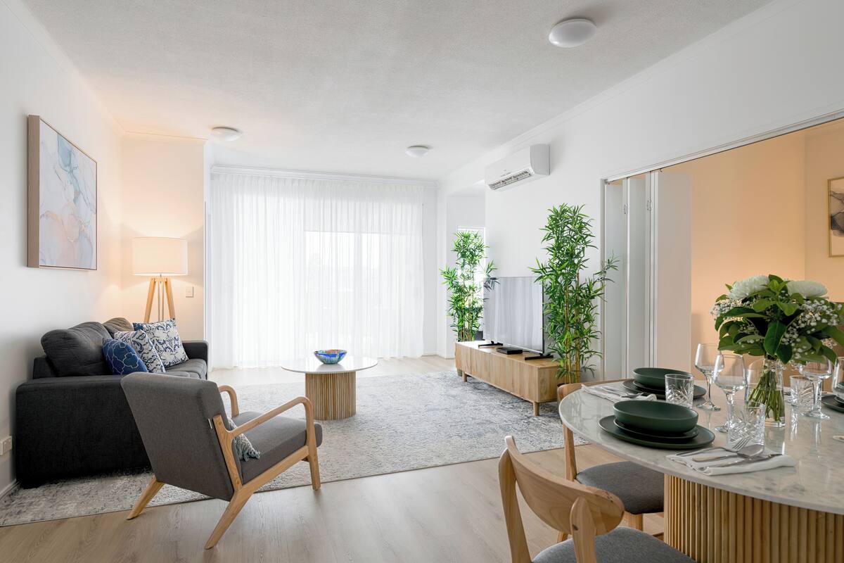 A modern living room with a sofa, chairs, a coffee table, a dining table set for four, and a large window with sheer curtains.