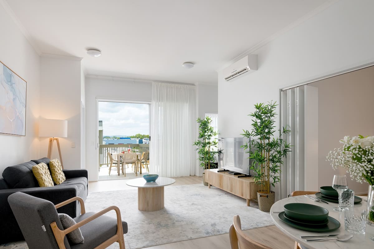 A modern living room with a sofa, coffee table, dining table, and a TV. There are plants and a view of the outdoors.