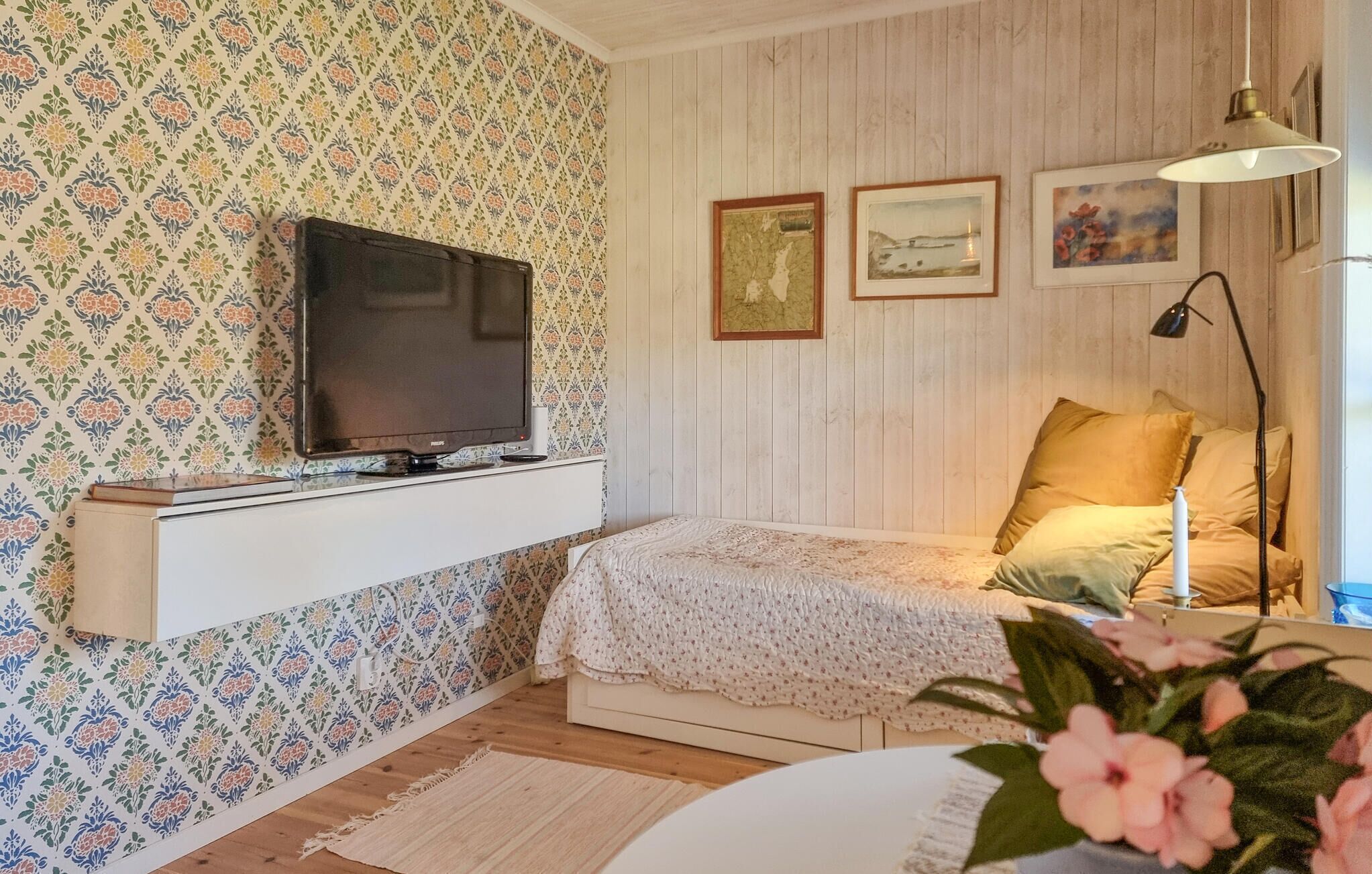 A cozy bedroom with a television on a shelf, a bed with a floral bedspread, and a window with a view of the countryside.