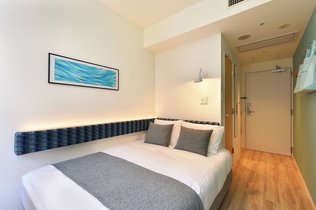 A modern hotel room with a neatly made bed, a wall-mounted artwork, a bedside lamp, and a door leading to another room.