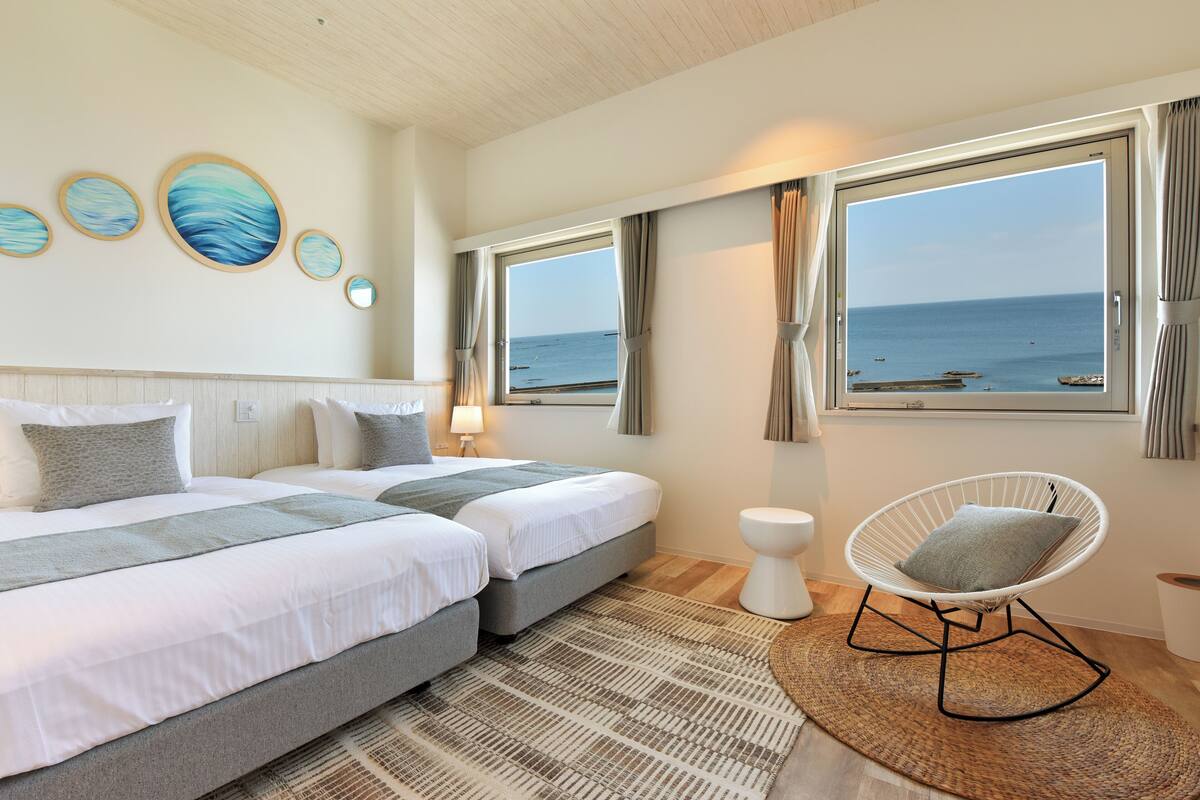 A hotel room with two beds, a round chair, a view of the sea, and artwork on the wall.