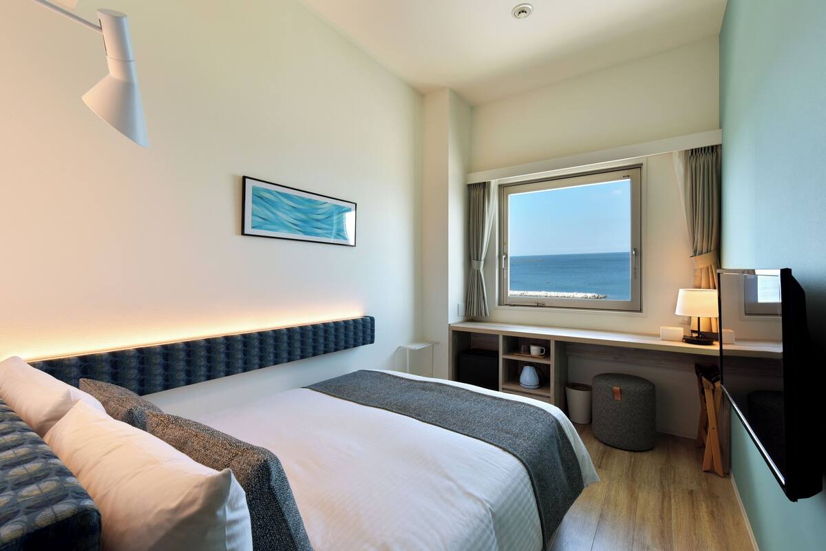 A hotel room with a bed, a desk with a lamp, a television, a window with a view of the sea, and a waste bin.