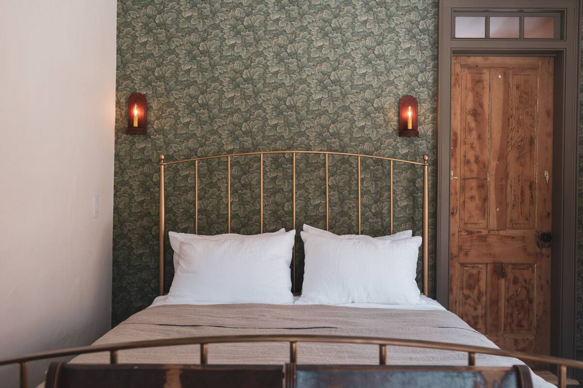A bed with a metal headboard, white pillows, and a wooden headboard. There are two wall-mounted lights and a wooden door in the background.