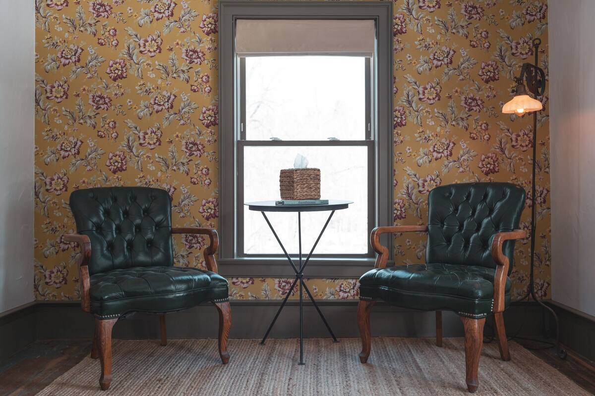 A room with two green leather chairs, a small table with a tissue box, and a window with floral wallpaper.
