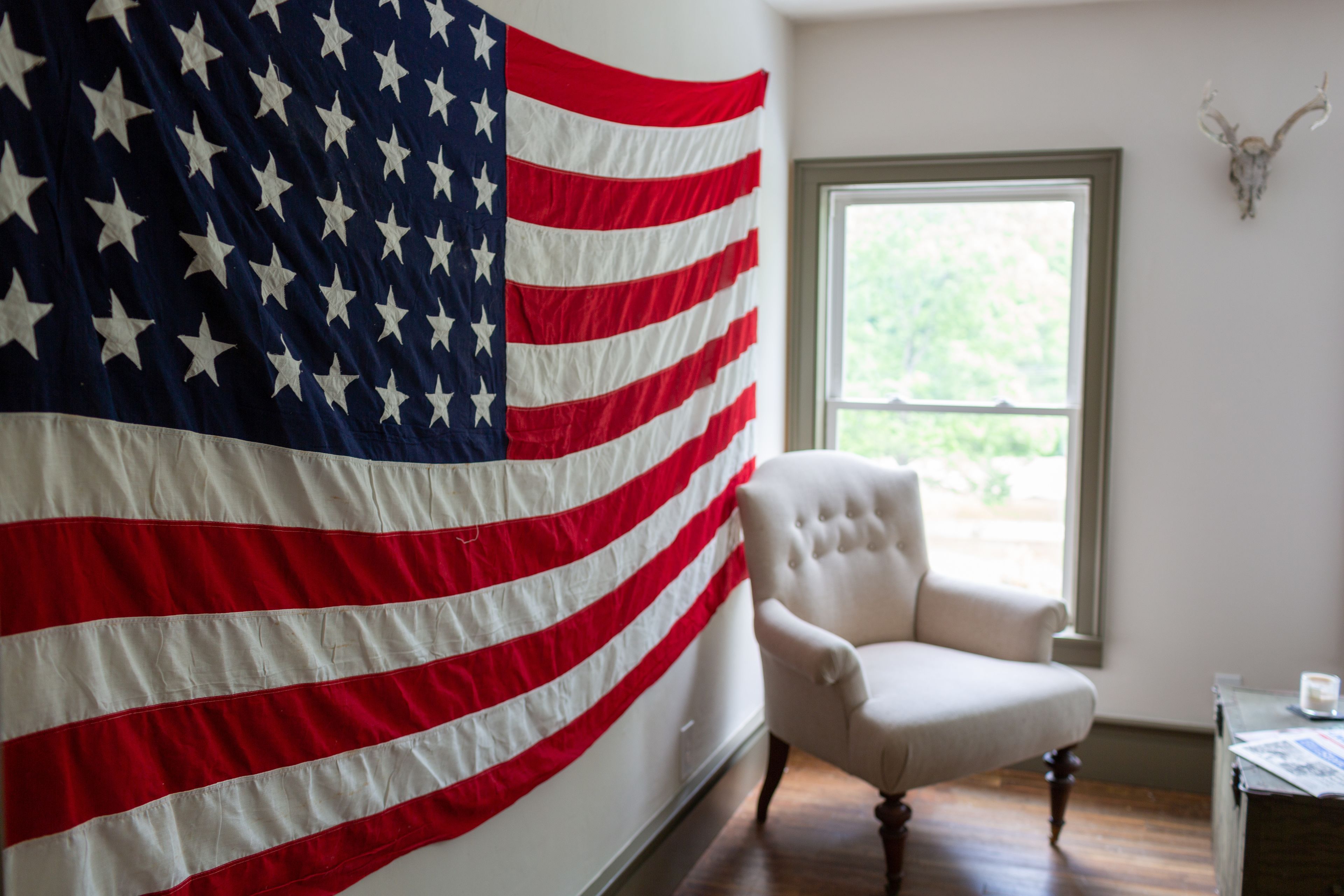 Una habitación con una gran bandera estadounidense, un sillón blanco, piso de madera, una ventana con vista a follaje y un cráneo de ciervo montado en la pared.