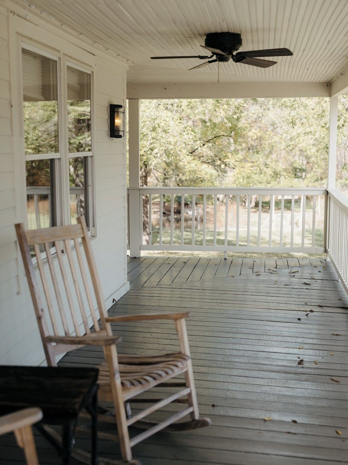 A covered porch with a ceiling fan, wooden chairs, and a view of trees.