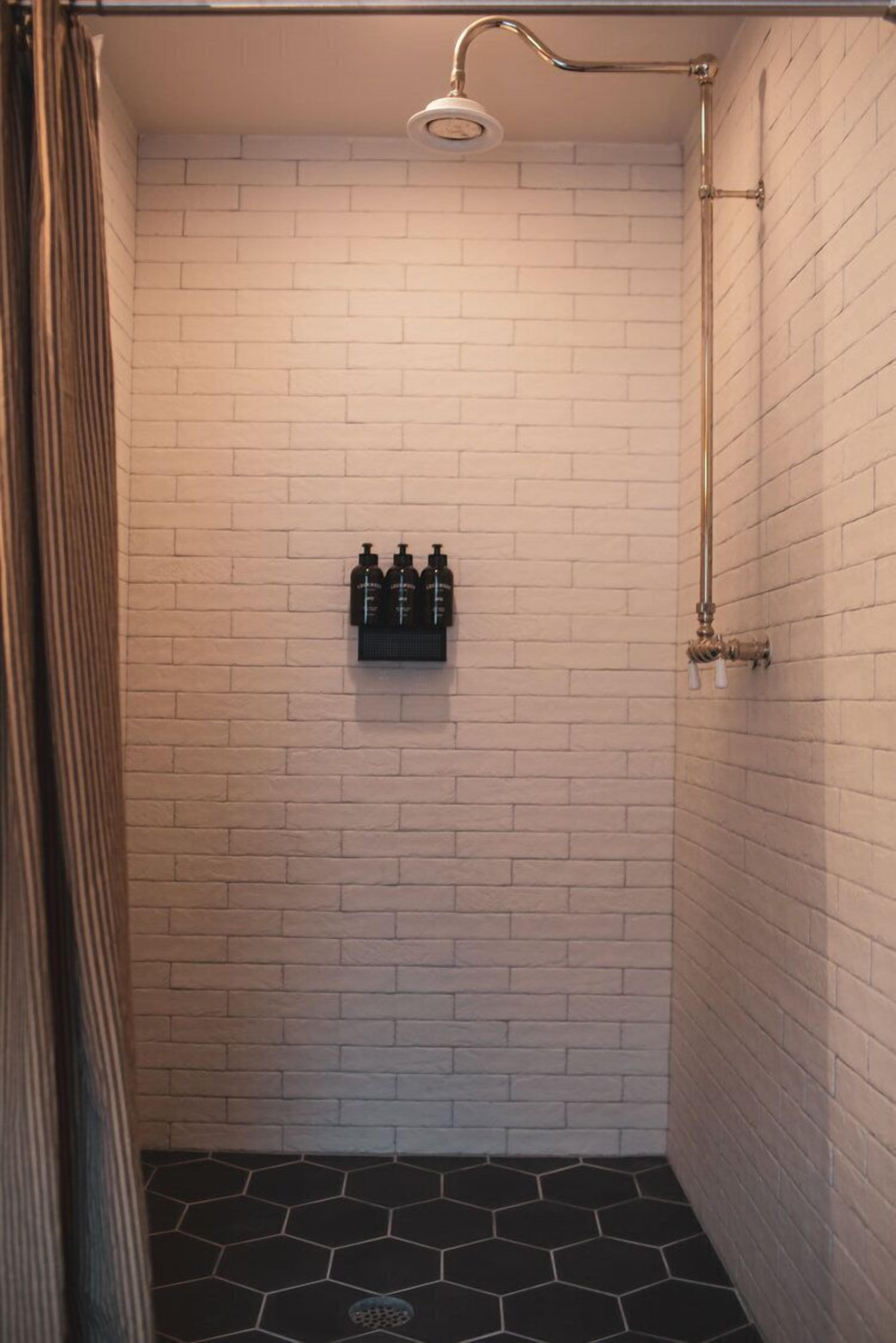 Ducha con pared de ladrillo blanco, piso de baldosas negras y repisa con botellas.