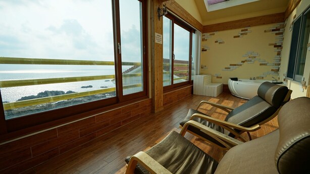 A room with wooden flooring, large windows with a view of the sea, and a massage chair. - Sunset in Garden (Seogwipo)