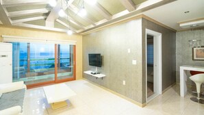 A spacious living room with a large sliding glass door leading to a balcony, a flat-screen TV on a stand, and a dining area with a white table and red chair. - Sunset in Garden (Seogwipo)