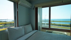 A room with a bed, a window with a view of the sea and a bridge, and an air conditioning unit. - Sunset in Garden (Seogwipo)