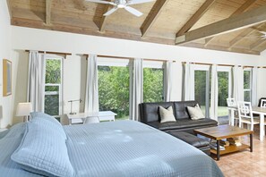 A spacious room with a large bed, a leather sofa, a dining table, and multiple windows offering a view of greenery.