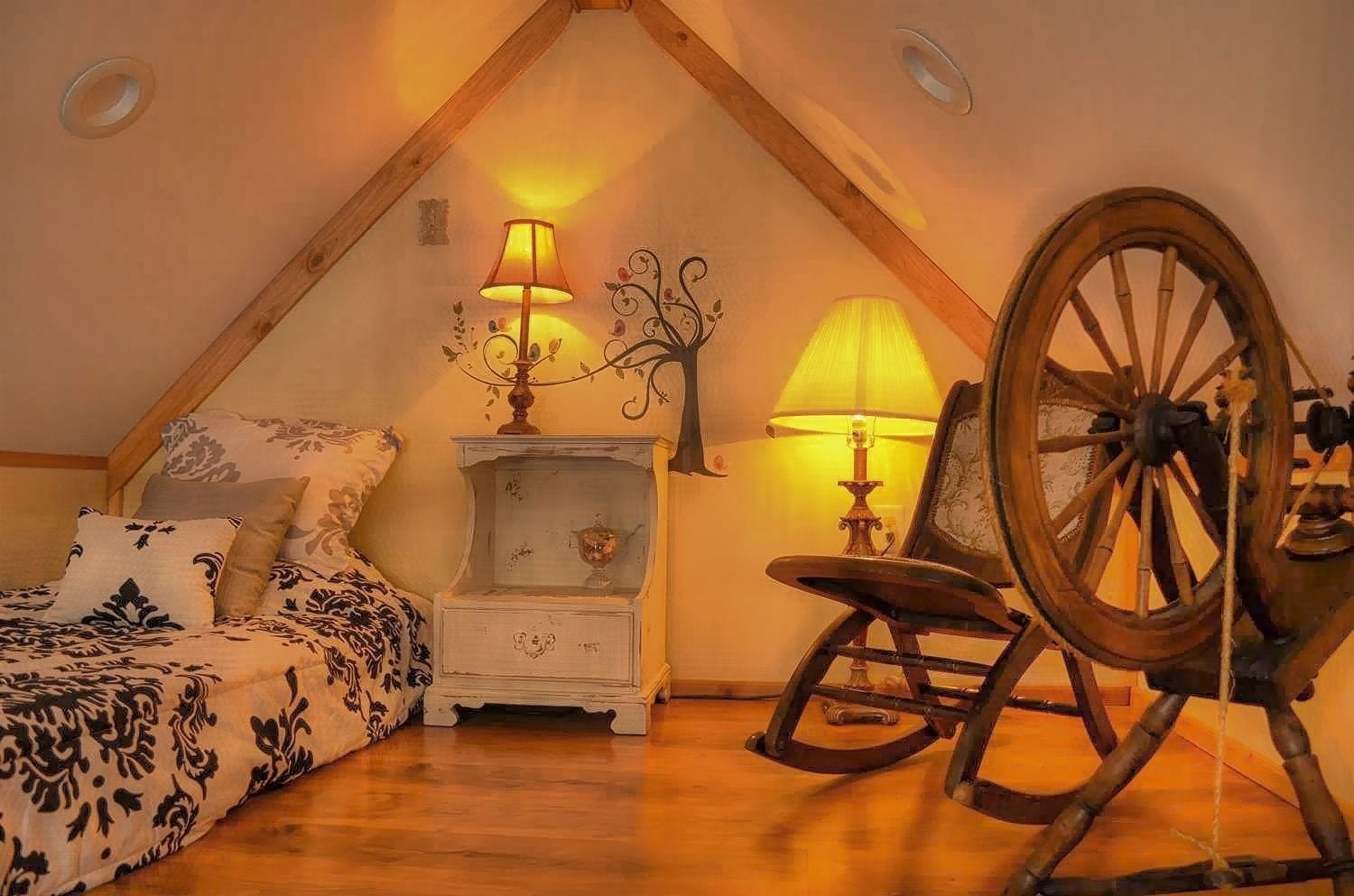 A cozy attic bedroom with a sloped ceiling, a bed with patterned bedding, a nightstand with a lamp, a rocking chair, and a large wooden wheel.