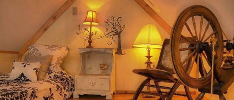A cozy attic bedroom with a sloped ceiling, a bed with patterned bedding, a nightstand with a lamp, a rocking chair, and a large wooden wheel.