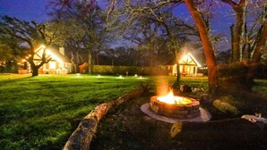 Une scène nocturne en plein air, avec un foyer, deux bâtiments illuminés et une pelouse bien entretenue.