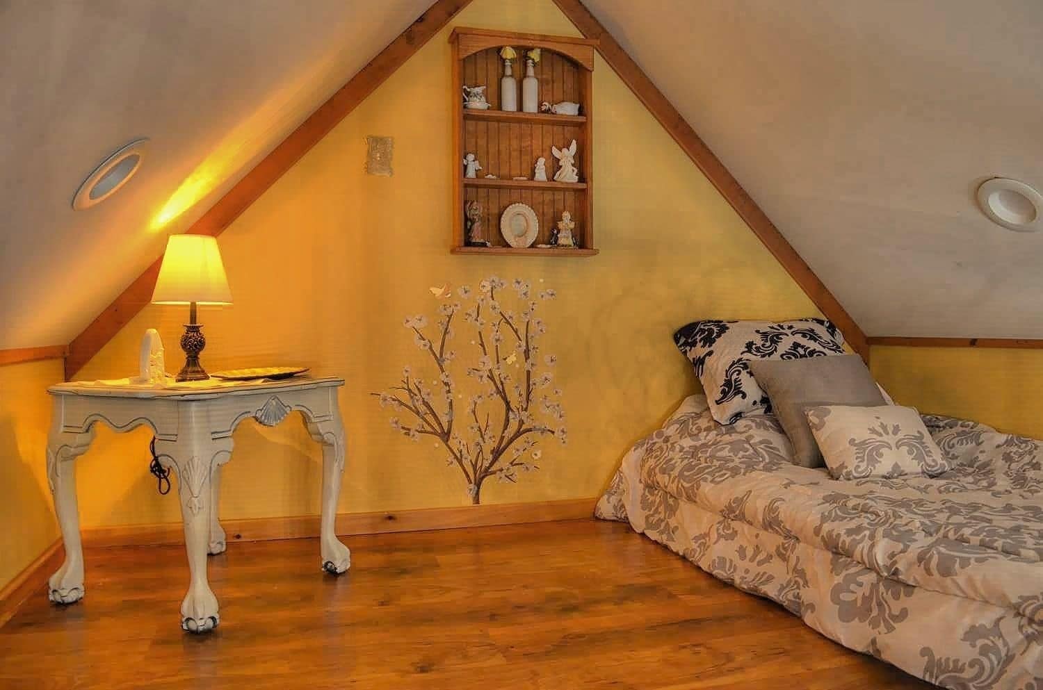 Ein gemütliches Schlafzimmer im Dachgeschoss mit geneigter Decke, einem Holzbett mit gemusterten Betttextilien, einem kleinen weißen Tisch mit einer Lampe und einem Regal mit Dekorationen.