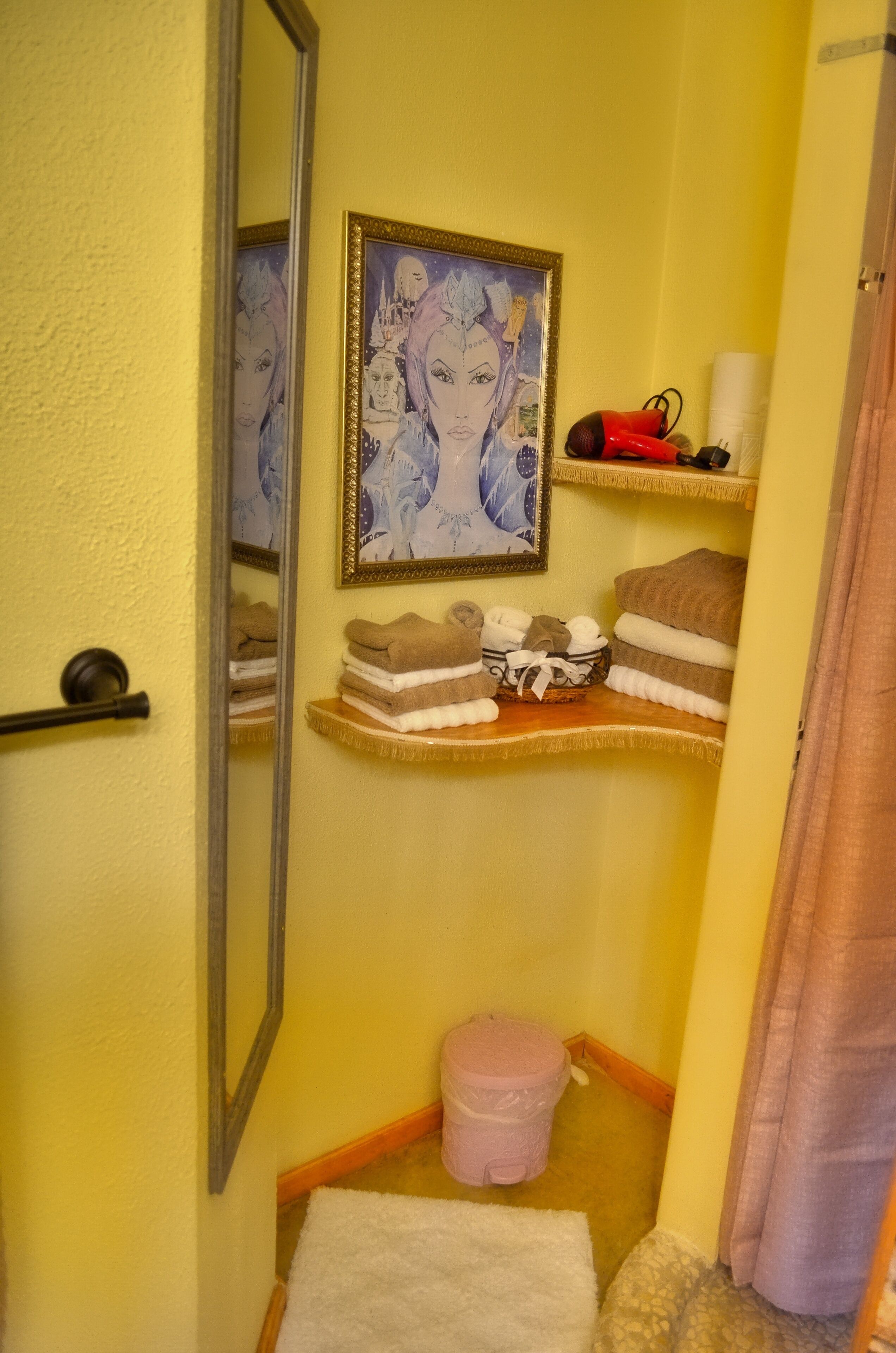 Une salle de bain avec un mur jaune, une œuvre d’art encadrée, un porte-serviettes avec des serviettes pliées, une poubelle rose et une serviette blanche sur le sol.